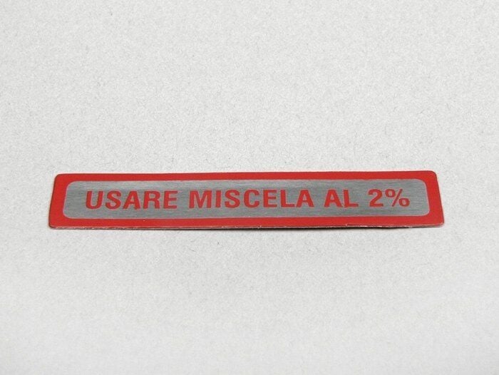 Tarra Vespan tankkiin, Usare Miscela al 2%, punainen Tarra Vespan tankkiin, Usare Miscela al 2%. Pituus 55mm, korkeus 10mm, väri punainen, pohja hopea. Tarra Vespan tankkiin, Usare Miscela al 2%, punainen